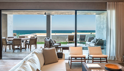 Minimalist indoor-outdoor living space with sliding glass walls opening to a furnished terrace and unobstructed ocean views.