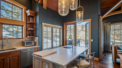 Stylish mountain home interior featuring a vaulted wood ceiling with exposed beams, large arched windows with natural light, a modern black dining table with brown and white chairs, and a cozy living area with a fireplace, gray sofas, and a wall-mounted TV.
