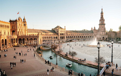 People walk near the fountain and canal of Plaza de España in Seville, Spain, a fun vacation spot for kids.