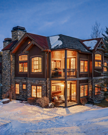 Close-up of a luxury mountain home with stone exterior, warm interior lights, covered balconies, and snow on the roof at twilight.