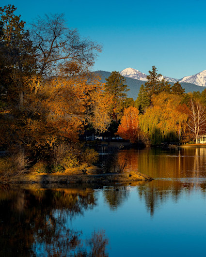 A serene autumn landscape in Bend, Oregon, captures vibrant fall foliage reflecting on calm water, with cozy mountain homes set against a backdrop of snow-capped peaks. This visually encapsulates the allure of autumn hiking and the luxury mountain retreat experience.