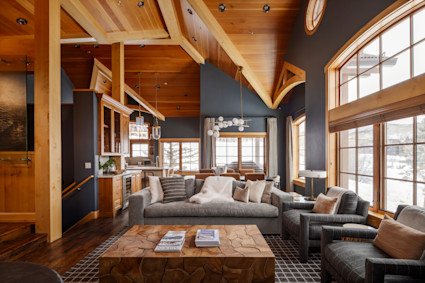 Open-concept luxury mountain great room with vaulted wood-beam ceilings, oversized windows, modern gray sofa and chairs, and a wet bar overlooking snowy views.