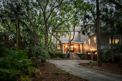 Lowcountry coastal home glowing at dusk, framed by towering live oaks and palmetto palms along a curved driveway.