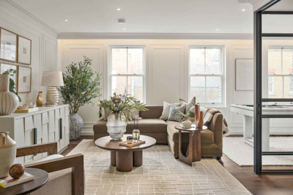 Bright, neutral-toned living room with two tall windows, an L-shaped tan sofa with pillows and a throw, a round wood coffee table with decor.