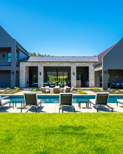 Modern dark gray estate with a stone-pillared patio, swimming pool, and lounge chairs under a clear blue sky.