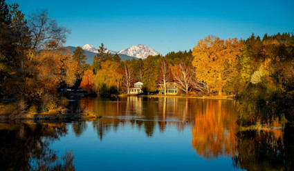 View of lake during fall with autumn-colored trees lining the waterfront and snowy mountains in the distance.