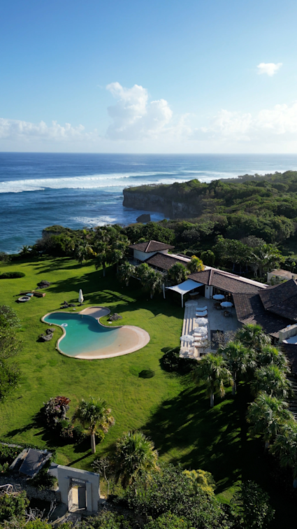 Aerial view of coastal estate with lagoon-style pool, terracotta roofs and lush tropical landscaping overlooking ocean cliffs