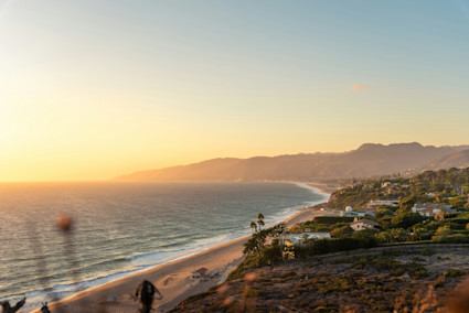 Golden-hour view of the Malibu coastline, waves rolling onto sandy beach below hillside homes and distant mountains.