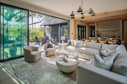 Airy open-plan living room with exposed wood beams, curved neutral furnishings, and sliding glass walls opening to a lush outdoor patio.
