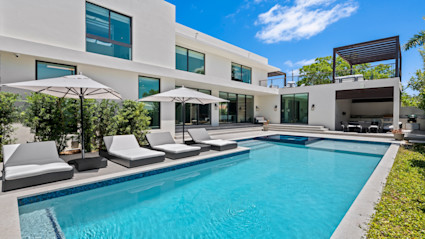 View of pool area with pool and hot tub with poolside lounge chairs and umbrellas with a small covered area and the rear exterior of a luxury home.