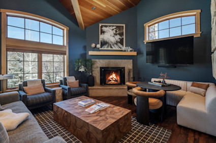 Cozy luxury mountain living room with vaulted wood ceiling, large arched windows, modern seating, and a lit stone fireplace overlooking snowy trees.