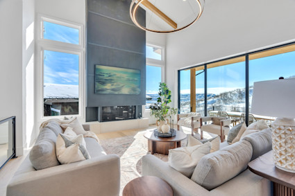 Sun-filled living room with double-height ceilings, a modern fireplace, and floor-to-ceiling glass framing snow-covered mountain views.