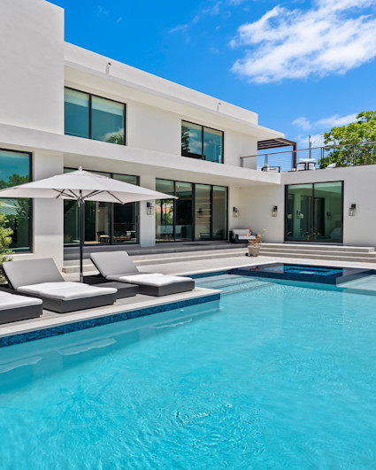 View of pool area with pool and hot tub with poolside lounge chairs and umbrellas with a small covered area and the rear exterior of a luxury home.