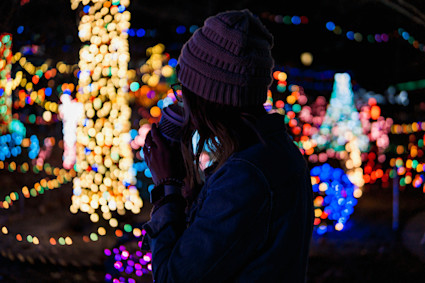 A woman gazes and colorful holiday lights while sipping a warm beverage.