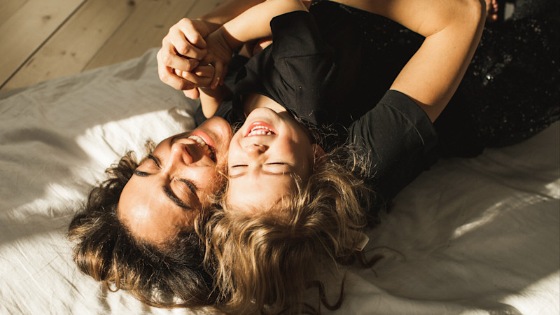Två personer i svarta kläder ligger på en säng i solljus, skrattar och håller varandras händer.