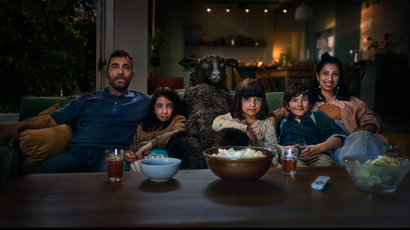 En familj sitter i en soffa med fåret Frank mellan sig medan de tittar på TV med popcorn och drycker på bordet framför.