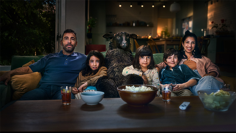 En familj sitter i en soffa med fåret Frank mellan sig medan de tittar på TV med popcorn och drycker på bordet framför.