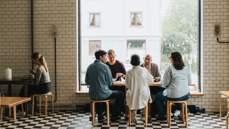 En grupp personer sitter vid ett bord på restaurang med ett stort fönster i bakgrunden. Bredvid dem sitter en ensam kvinna vid ett bord.