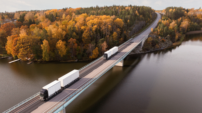 Två lastbilar kör på en bro över en sjö med skog med höstlöv i bakgrunden.
