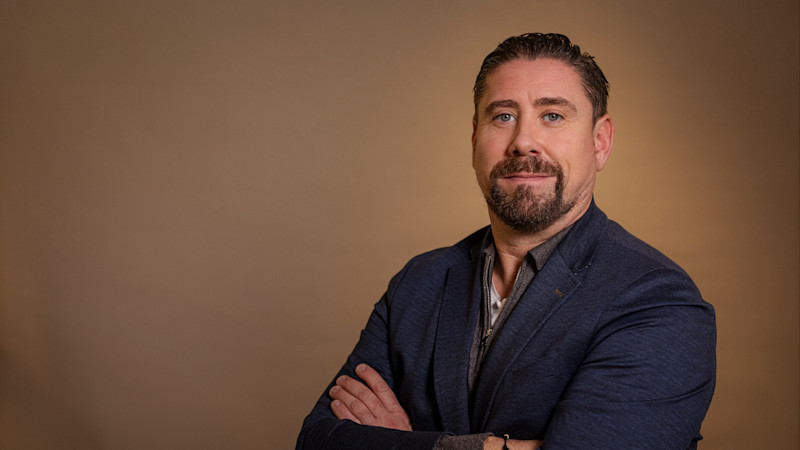 En person i mörkblå kavaj med armarna korsade mot en varm brun bakgrund, med ett vänligt leende.