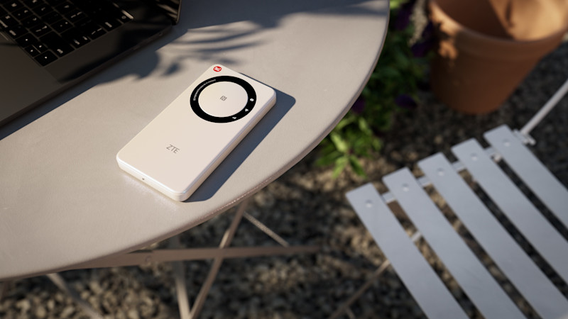 Portabel 5G-router från ZTE ligger på ett runt utebord i metall, bredvid en bärbar dator. Bordet står på grus, och i bakgrunden syns en vit trädgårdsstol och en blomkruka i terrakotta.