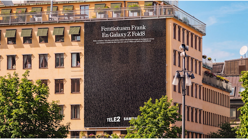 Stor reklamaffisch på husvägg i stad med texten "Femtiotusen Frank, En Galaxy Z Fold8" för Tele2 och Samsung.