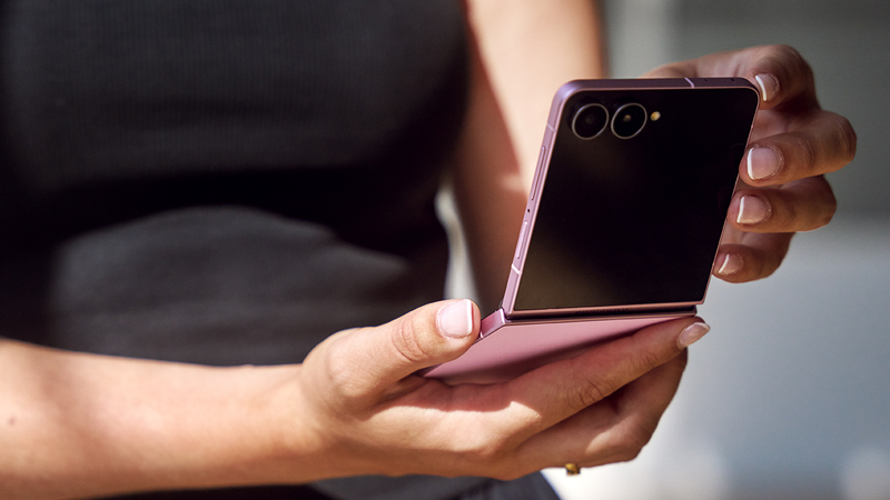 Närbild på händer som håller en uppvikt smartphone. Ena halvan visar en mörk skärm med två kameror, medan andra halvan visar en rosa baksida.