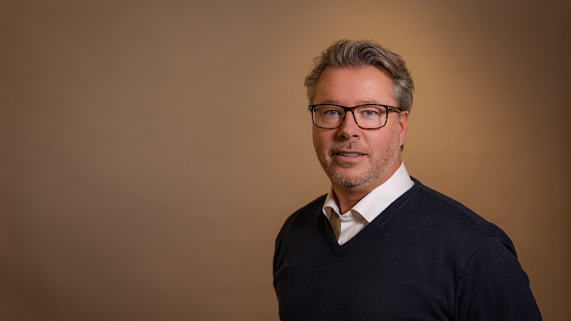 En person med glasögon i mörkblå tröja över vit skjorta mot en varm beige bakgrund.