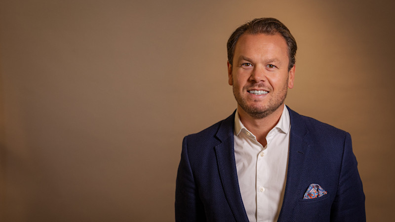 En leende person i mörkblå kavaj med mönstrad näsduk och vit skjorta mot en varm brun bakgrund.