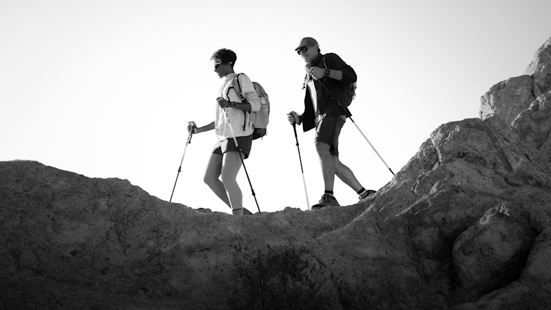 Två vandrare med ryggsäckar och vandringsstavar går längs en klippig stig i svartvitt fotografi.