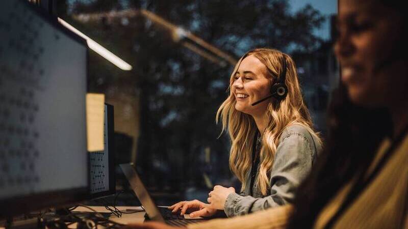 Två kvinnor sitter framför datorskärmar på ett kontor. De ler och bär headset.