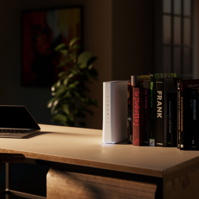En rad böcker, en vit bredbandsrouter och en bärbar dator på ett skrivbord i ett dunkelt rum med en krukväxt och konst i bakgrunden.