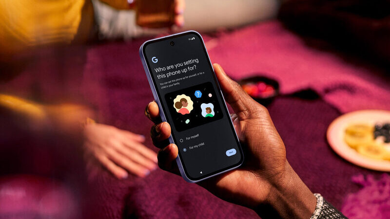 Närbild på en hand som håller i en Google Pixel-mobil. I bakgrunden skymtar en annan person samt en röd bordsduk.