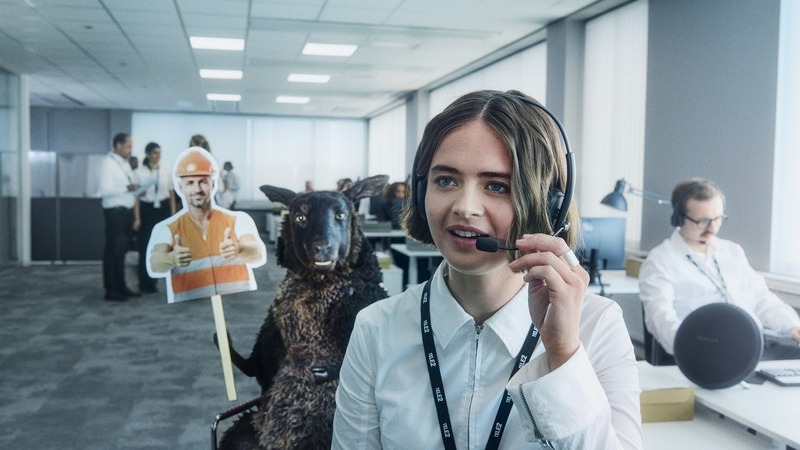 Ett kontorslandskap med personer i vita skjortor och mörka byxor. Längst fram en kvinna med headset och bakom hemme står det svarta fåret Frank som håller i en skylt med en man i orange skyddsväst och hjälm.