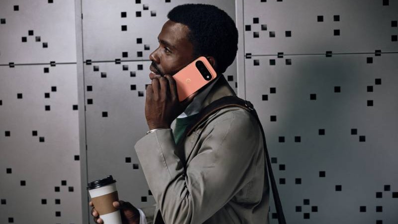 Profilbild av man som står vid en grå vägg och pratar i en rosa smartphone med kamera högst upp på baksidan. Mannen bär kavaj och håller i en kaffemugg.