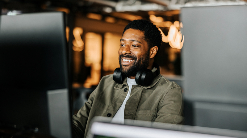 En man sitter framför en datorskärm på kontor med hörlurar runt halsen.