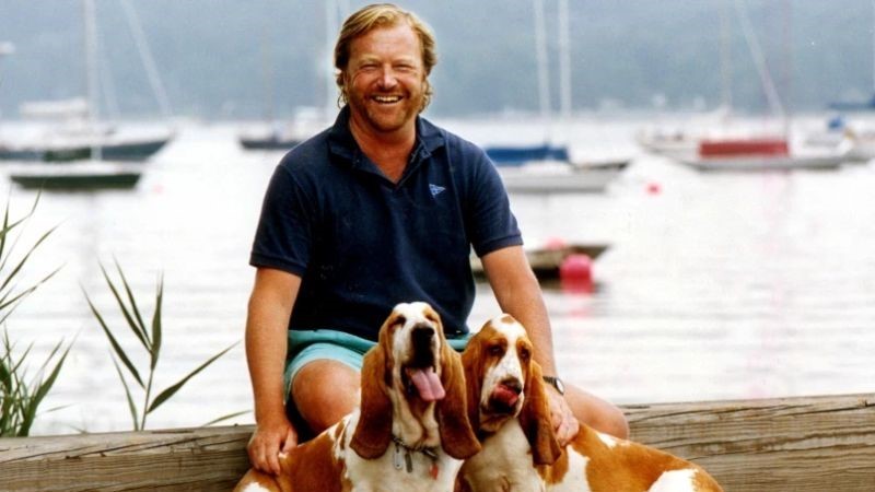 Affärsmannen Jan Stenbeck som grundade bland annat Tele2 sitter på en brygga vid vattnet klädd i shorts och pikétröja. Framför honom sitter två basset-hundar och i bakgrunden syns vatten och segelbåtar. Han ler och ser avslappnad ut.