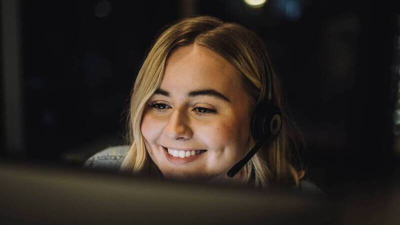 Närbild på blond kvinna med headset framför en datorskärm.