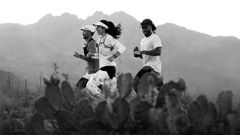 Tre löpare springer i ökenterräng med kaktus i förgrunden och berg i bakgrunden, svartvit bild.