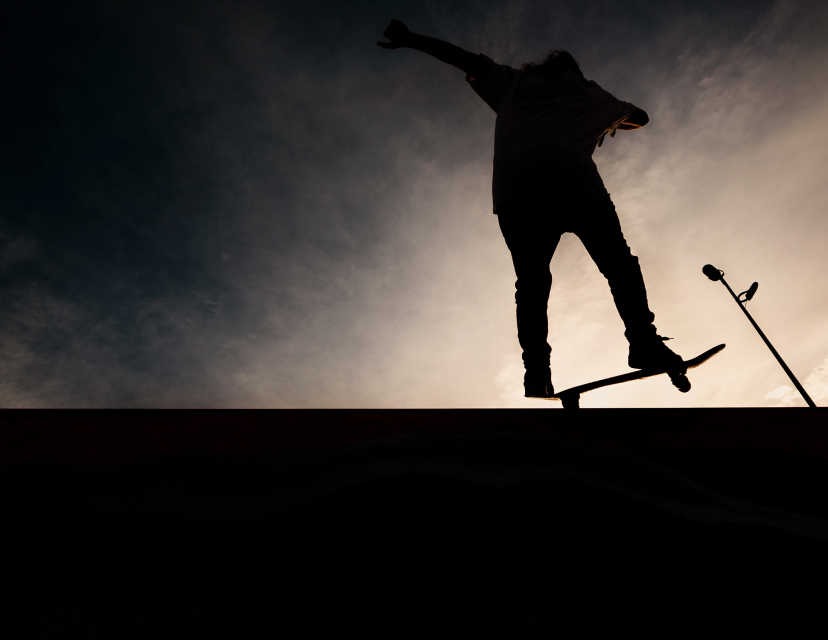 Silhuett av en skateboardåkare som hoppar mot en dramatisk molnig skymningshimmel, med en lyktstolpe i bakgrunden.