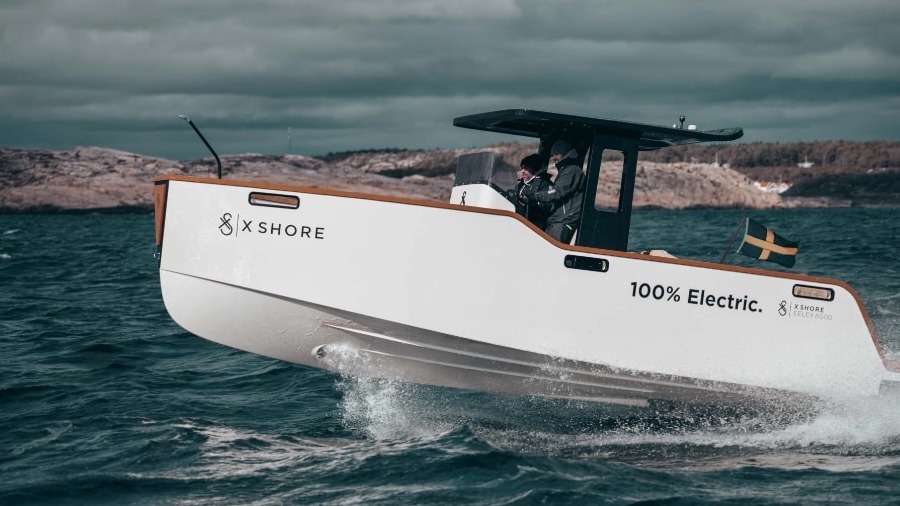 X-shores elektriska båt åker i hög hastighet över havet med en klippig ö i bakgrunden. Båten är liten och vit med mörkt tak och körs av en person.