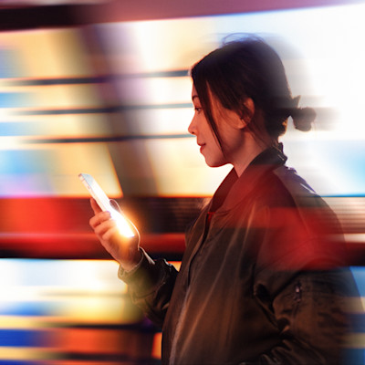 Profil av person i läderjacka som tittar på en lysande mobiltelefon mot en suddig bakgrund med färgglada ljus.