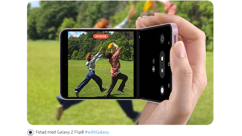 En hand håller en lila Samsung Galaxy Z Flip som filmar två personer som leker med en frisbee på en grön gräsmatta.