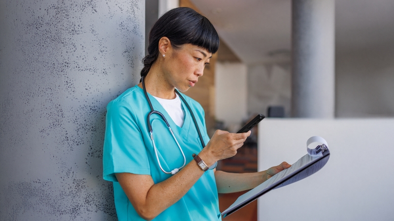 En kvinna i sjukvårdskläder med stetoskop runt halsen håller i en mobil och läser ur ett anteckningsblock.
