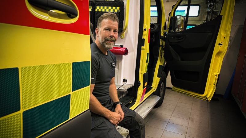 Joakim Sihlberg sitter i dörren till en ambulans i ett garage, klädd i ambulansuniform. Han har skägg och brunt hår och ler mot kameran.