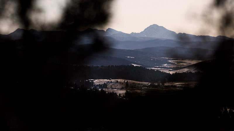 Utsikt genom mörka trädsilhuetter mot ett dalgångslandskap med skogar och berg i bakgrunden under mulen himmel.