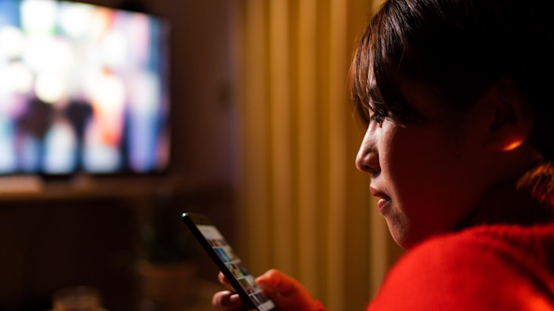 En ung kvinna klädd i röd tröja tittar på en mobiltelefon. Hon har mörkbrunt hår med lugg. Bakom henne skymtar en vägg som är randig i gulbruna nyanser samt en tv-skärm med suddigt innehåll.