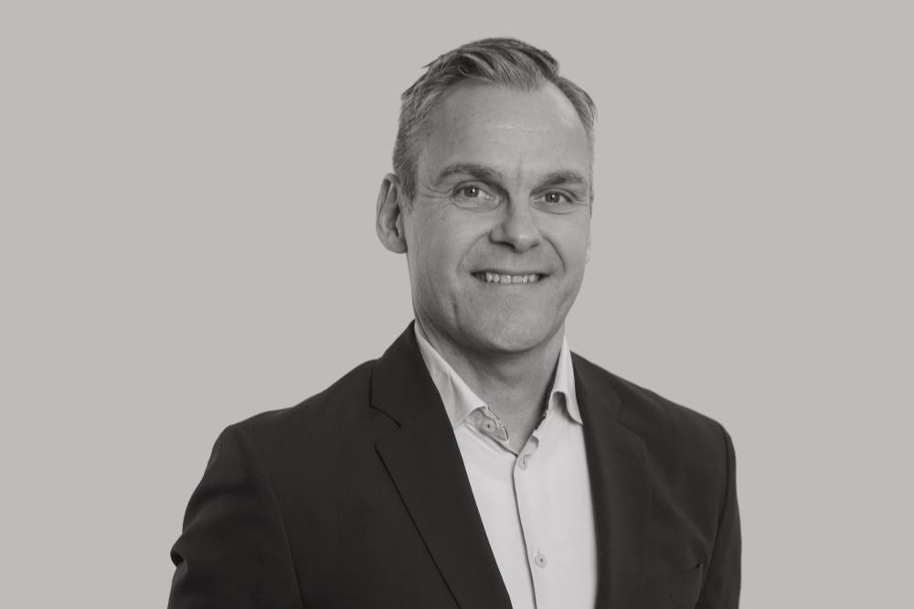Svartvit porträttbild av Michel Garcia på Instalco. Han har kort ljusbrunt hår och ler. Han är klädd i mörk kavaj och vit skjorta och står framför en grå vägg.