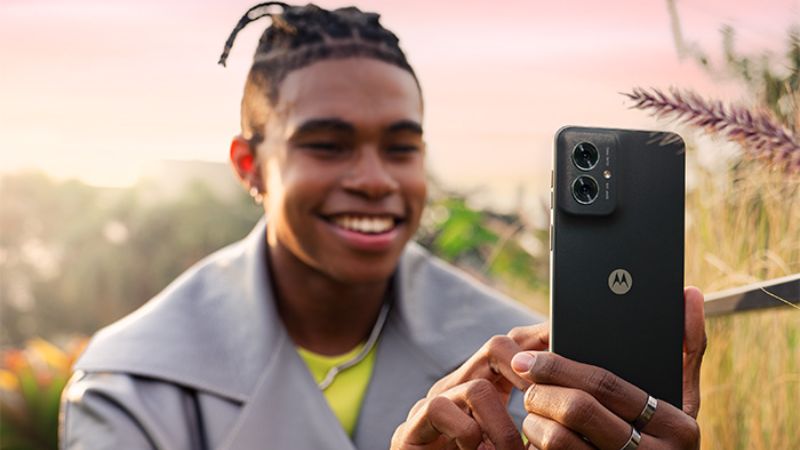 Ung man fotograferar med en svart Motorola moto G55-mobil. Han ler och är klädd i grå jacka. I bakgrunden syns buskar och solsken.