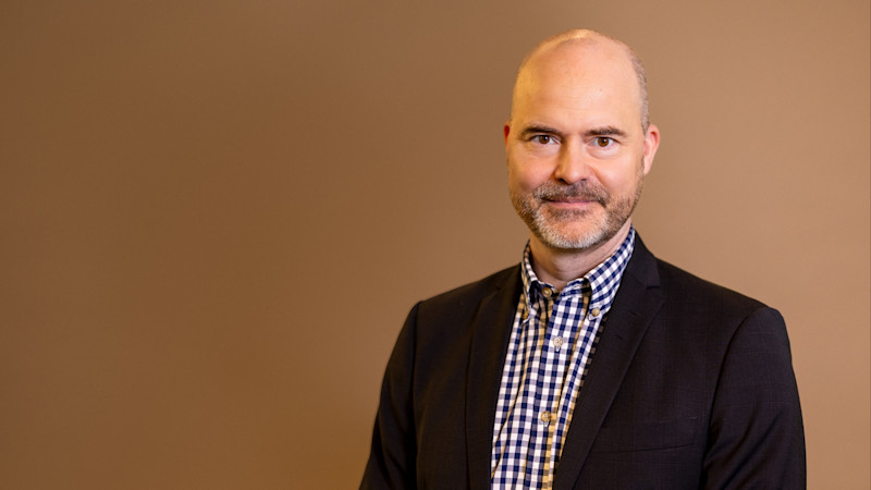 En man i mörk kavaj och rutig skjorta mot en brun bakgrund, med skägg och skallig hjässa.